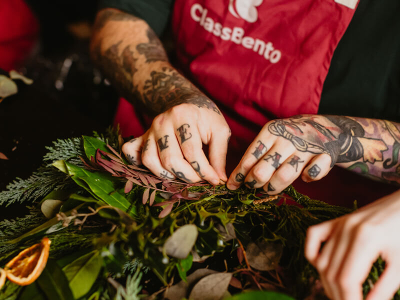 A close up of two hands decorated with tattoos making a Christmas wreath from greenery